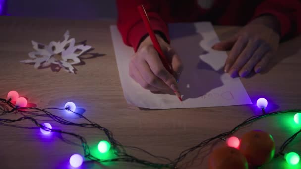Winter holidays, female hands with a red pencil write a letter with wishes for Santa Claus while sitting at table with lights in a cozy dark room — стоковое видео