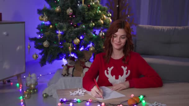 Letter to Santa Claus, happy dreamy girl writes a wish list for the new year and then lays down note near the christmas tree in a cozy dark room with a festive interior — Stok video