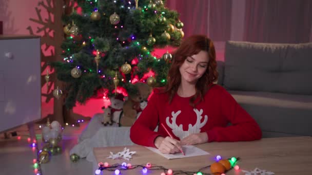 Girl writing xmas letter to Santa Claus, dreamy young woman writing wish list for new year on background of Christmas tree at cozy dark living room with festive interior — ストック動画