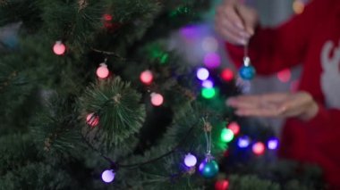new year celebration, girl in red sweater decorating christmas tree with balls and bells, lights on artificial branches close-up