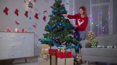 happy girl in red sweater with a deer decorates a christmas tree with a lights garland at home, lots of gift boxes under the tree