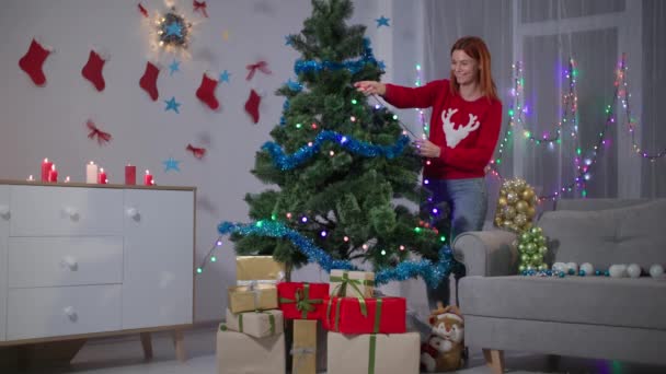 Happy girl in red sweater with a deer decorates a christmas tree with a lights garland at home, lots of gift boxes under the tree — Stok video