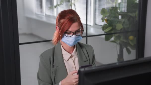 Kwarantanna, młoda kobieta w okularach z maską medyczną na twarzy pracuje przy komputerze i używa smartfona siedzącego przy stole w biurze, patrzącego w kamerę — Wideo stockowe