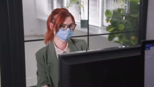 Pandemie, een jonge vrouw met een bril voor het zien in een medisch masker op haar gezicht ter bescherming tegen een virus en infectie praat aan de telefoon terwijl ze achter de computer op kantoor zit — Stockvideo