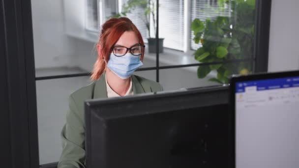 Kantoor coronavirus, vrouwelijke werknemer met bril en een medisch masker op haar gezicht praat op een mobiele telefoon met computer en kijkt naar camera — Stockvideo