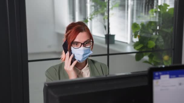 Gözlüklü ve yüzünde tıbbi maske olan genç bir kadın ofiste çalışıyor ve ofiste virüsün yayılmasını önlemek için önlemler alıyor, kameraya bakın. — Stok video