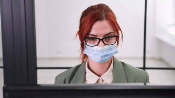 Portretten van vrouwelijke werknemer met bril en medisch masker te beschermen tegen virus en infectie werkt als computer, dan neemt haar bril af en kijkt naar de camera — Stockvideo