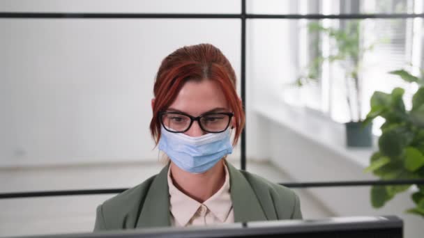 Jonge vrouw met een bril en een medisch masker om te beschermen tegen virussen en infecties werkt op de computer in het kantoor na het openen lockdown, glimlacht en kijkt naar de camera — Stockvideo