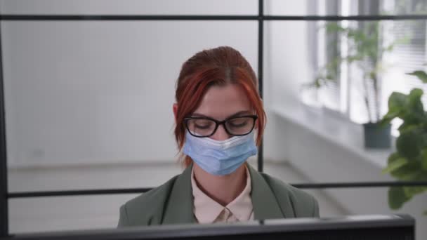 Pandemie werk, jonge vrouwelijke werknemer met een medisch masker op haar gezicht werkt in het kantoor op de computer, het observeren van moderne voorzorgsmaatregelen om de verspreiding van het virus te voorkomen, doet masker af, glimlacht en ziet er een — Stockvideo