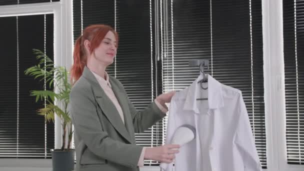 현대 과학 기술, 행복 한 미소를 짓는 여성은 창문을 등지고 방에 서 있는 동안뜨거운 증기로 손 증류기를 사용하여 셔츠를 다리미질한다 — 비디오