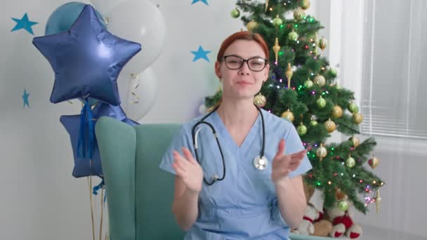 Felicitaciones en línea en feliz navidad, mujer joven médico en gafas y uniforme mira a la cámara y habla discurso de felicitación cerca del árbol de Navidad en la oficina — Vídeo de stock