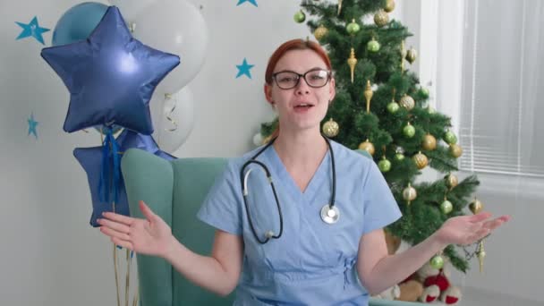 Online greeting happy new year, female doctor in eyeglasses and blue uniform looking at camera and talking congratulatory speech near christmas tree in office or at home — Stok video