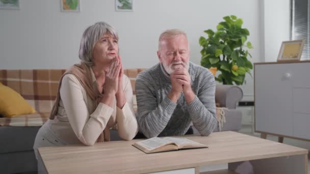 Elbűvölő idős férfi és nő csukott szemmel és összehajtott kézzel reménnyel a szívükben térdelnek és imádkoznak Istenhez az asztalnál a szobában. — Stock videók