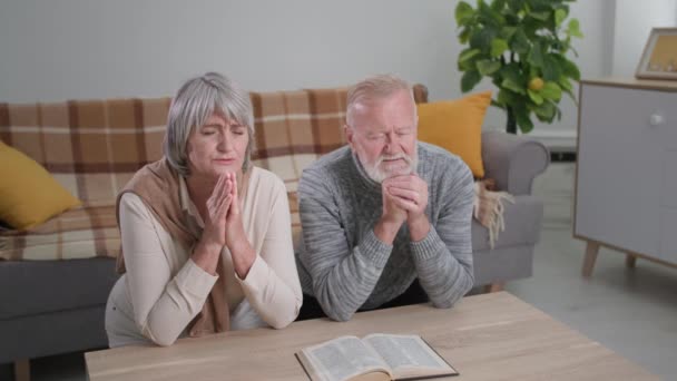 Oudere vrouw met haar oude man knielen met gevouwen handen en gesloten ogen en bidden tot God met hoop en geloof in hun hart, kijken naar de camera — Stockvideo