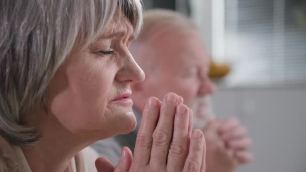 Geloof, een oudere vrouw met haar oude man bidden met gesloten ogen en gevouwen handen, staande op het rijtuig met beven en tederheid voor God in de kamer, close-up — Stockvideo