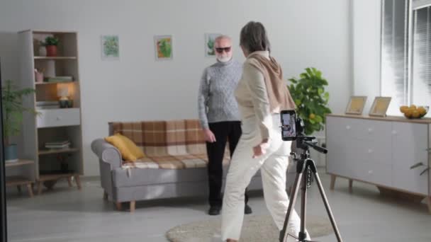 Vieux blogueurs, retraités homme et femme tournent joyeusement des danses sur une caméra smartphone pour les réseaux sociaux populaires dans la salle de séjour confortable, montrer la classe à la caméra — Video