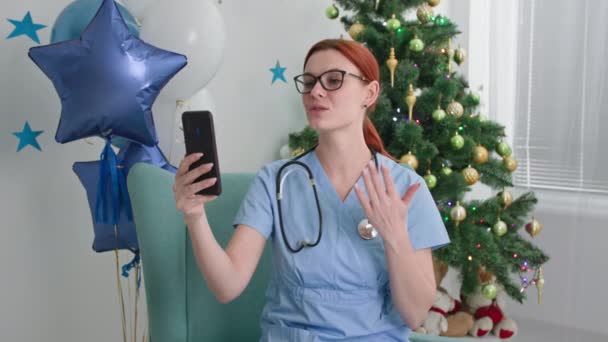 С Новым годом онлайн молодая женщина-врач с фонендоскопом общается с пациентами по телефону по видеосвязи, поздравляя с зимними праздниками в украшенном офисе с новогодней елкой — стоковое видео