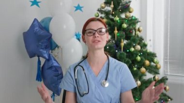 Gözlüklü genç bayan doktor, yılbaşı tatilinde çalışırken Noel ağacıyla ofiste otururken ailesini videoyla tebrik ediyor.