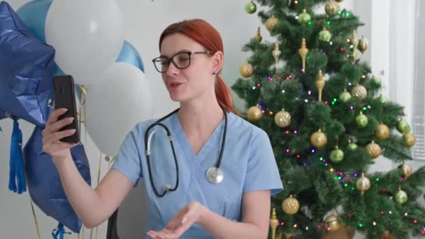 Holidays online in workplace, charming doctor congratulates his family on New Years on camera on smartphone while working on New Year Eve — Stock Video