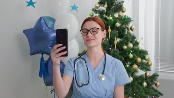 Navidad en línea, un encantador médico felicita a la fiesta por videollamada utilizando un teléfono inteligente mientras está sentado en la oficina sobre el fondo de un árbol de Navidad y globos — Vídeo de stock
