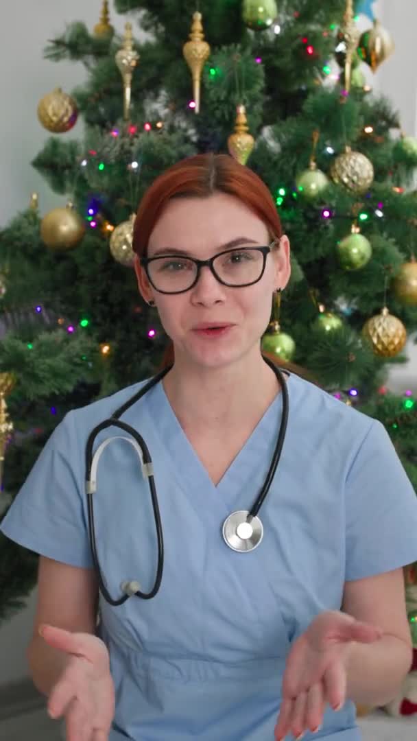 Médico felicita en las vacaciones en línea, las mujeres médico oficial felicita en las vacaciones de Navidad por enlace de vídeo sentado fondo del árbol de Navidad, pantalla vertical — Vídeo de stock
