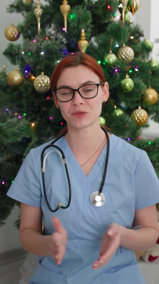 Nuevo año en línea, médico femenino felicita en las vacaciones de Navidad por enlace de vídeo sentado fondo del árbol de Navidad, pantalla vertical — Vídeos de Stock