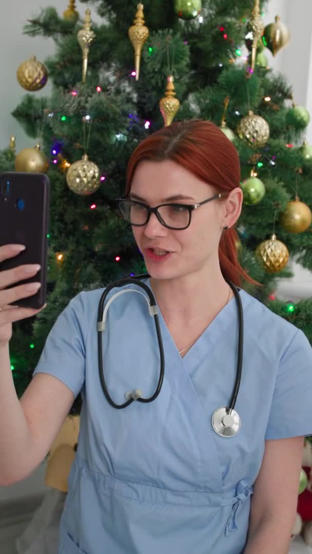 Fjärrgratulationer, söt kvinnlig läkare gratulerar online-patienter på videokamera med hjälp av smartphone när du sitter i ett kontor bakgrund av en julgran, vertikal skärm — Stockvideo