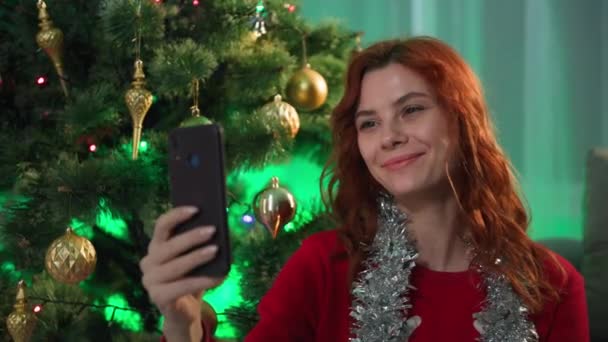Bayram interneti, neşeli yağmurla eğlenen genç bir kadın tatilde ve Noel atmosferinde yılbaşı ağacının yanında otururken akıllı telefondan arkadaşlarıyla ve akrabalarıyla görüntülü iletişim kuruyor. — Stok video