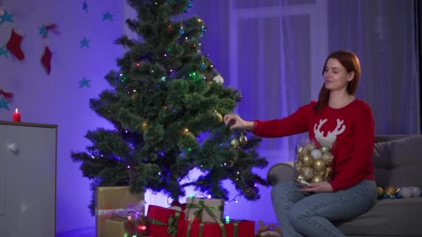 Winterurlaub, lächelnde Frau im Pullover mit Hirsch schmückt einen künstlichen Weihnachtsbaum mit einer Girlande aus festlichem Spielzeug in einem gemütlichen Raum — Stockvideo