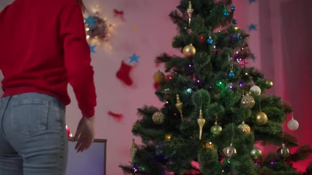 Voorbereiding op het nieuwe jaar, een jonge huisvrouw versiert een kunstmatige boom met een slinger van kerstballen tijdens de voorbereiding van de wintervakantie in een gezellige kamer — Stockvideo
