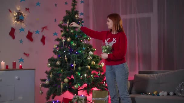 Während sie das Haus auf die Winterferien vorbereitet, schmückt eine süße Frau im roten Pullover mit Hirsch einen ökologischen Baum mit festlichem Spielzeug und einer Gorland in einem gemütlichen Raum im Licht — Stockvideo