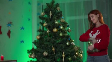 Mutlu Noeller. Çekici bir ev hanımı, sıcacık bir odada ışıkları olan güzel topları asan bir Noel ağacını süslemekten zevk alır.