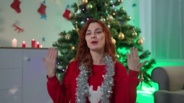 Video tebrikler, geyik ve dekoratif yağmurlu süveterli mutlu kadın Noel tatilinde ailenizi kutluyor. Yılbaşı ağacının arka planında festival odasındaki video bağlantısıyla oturuyor.