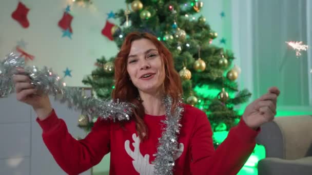 Saludos de Año Nuevo en línea, mujer encantadora con bengalas se comunica a través de videollamada con amigos y felicita en las vacaciones de Navidad sentado fondo del árbol de Navidad en una hermosa habitación — Vídeos de Stock