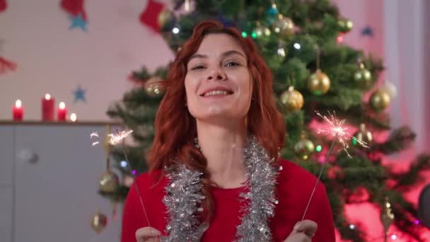 Charmante Frau mit Wunderkerzen spricht per Videoanruf und grüßt Neujahr, während sie zu Hause im Hintergrund des Weihnachtsbaums sitzt und in die Kamera schaut — Stockvideo