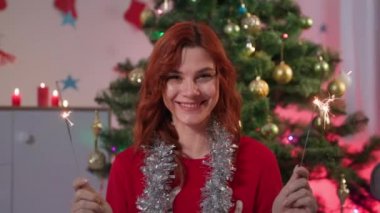 Elinde maytap olan gülümseyen kadın portresi dekorasyon odasındaki yeni yıl ağacının arka planında oturuyor. Işıklar doğum günlerini kutlamak için, kameraya bakıyor.