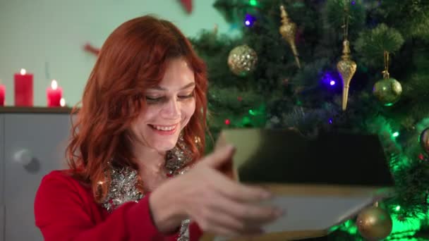 Nieuwjaarsgeschenken, jonge glimlachende vrouw verheugt zich over de verrassing terwijl ze zit in de buurt van een kerstboom in een mooie kamer — Stockvideo