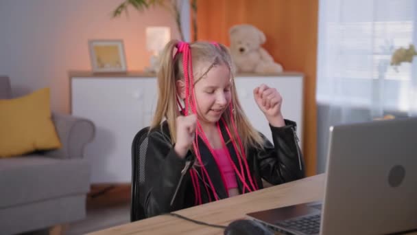 Adicción al juego, una linda chica con trenzas de colores disfruta jugando en un ordenador portátil mientras está sentado en una mesa en una habitación acogedora — Vídeo de stock