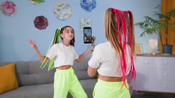 Hobbies modernos, uma linda criança do sexo feminino com uma namorada filmando conteúdo de vídeo de dança para redes sociais usando um estabilizador para um smartphone na sala de estar — Vídeo de Stock