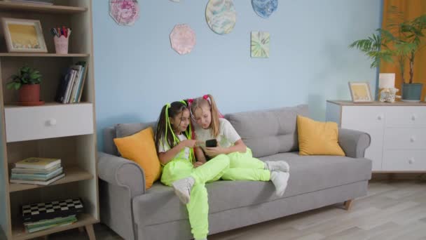 Τα παιδιά ξεκουράζονται στο σπίτι, αξιολάτρευτο έφηβα κορίτσια με πανομοιότυπα κοστούμια και κομψό χτενίσματα διασκεδάζουν βλέποντας δημοφιλή κοινωνικά δίκτυα χρησιμοποιώντας ένα smartphone, ενώ κάθεται στον καναπέ στο δωμάτιο — Αρχείο Βίντεο