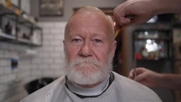 Мужской салон, портрет стильного старика с седой бородой, делающего стрижку и укладку в парикмахерской, сидящего в кресле парикмахера в помещении — стоковое видео