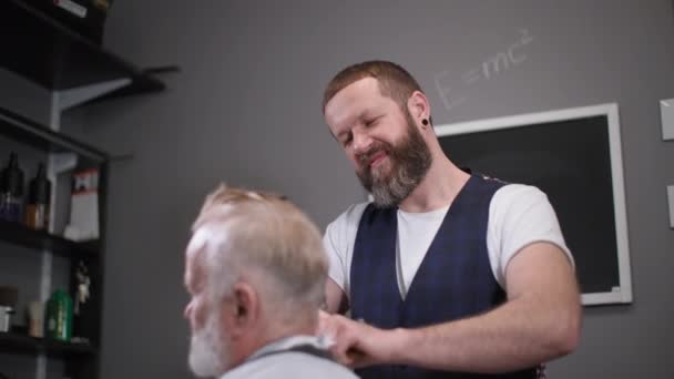 Professionale taglio di capelli maschile dà un taglio di capelli con un pettine e trimmer ad un elegante pensionato maschile in un popolare barbiere — Video Stock