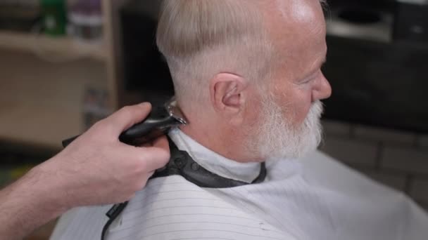 Мужской салон, пожилой мужчина с бородой делает современную стрижку с триммером в модной парикмахерской — стоковое видео