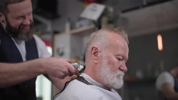 Sorridente cortador de cabelo masculino faz um aparador para um cliente idoso com barba em uma barbearia — Vídeo de Stock