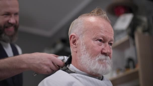 Barbiere, sorridente uomo anziano con la barba fa un taglio di capelli utilizzando un trimmer da un parrucchiere moderno in un salone da uomo — Video Stock