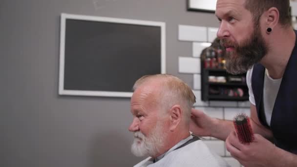 Encantador anciano en silla de peluquero hace un corte de pelo y peinado en un peluquero popular en el salón de los hombres — Vídeo de stock