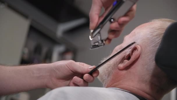 Парикмахер-мужчина делает стрижку и укладку старику с бородой, используя триммер и расческу в парикмахерской, крупным планом — стоковое видео