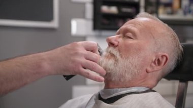 Gri saçlı ve sakallı klas bir müşteri, kuaförde modern bir berber kullanarak bıyığını kesiyor.
