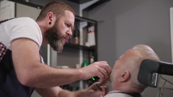 Professionale barbiere moderno fa un taglio di capelli e lo stile di baffi e barba per un uomo anziano elegante utilizzando un trimmer nel salone — Video Stock