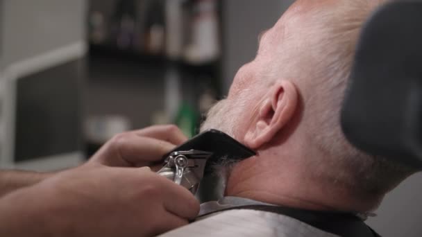 Barbiere professionista fa uno stile barba e taglio di capelli con un trimmer e pettine a un uomo anziano in un barbiere — Video Stock